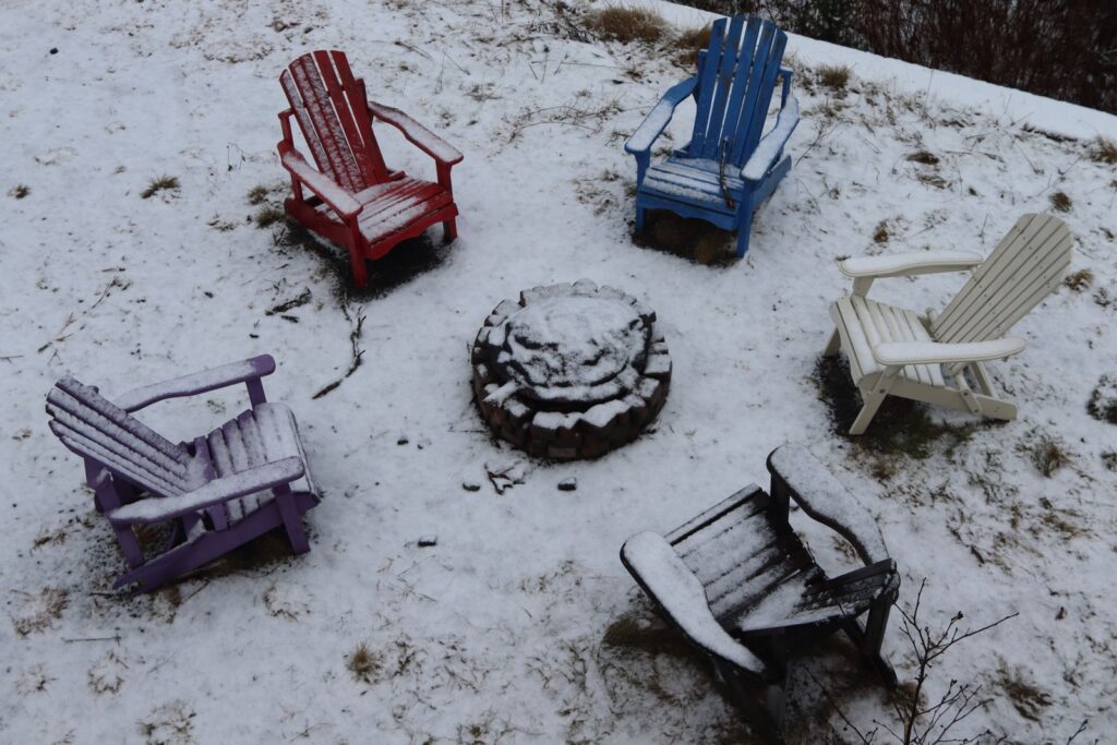 Five colorful Adirondack chairs in a circle around a stone fire pit in light snow