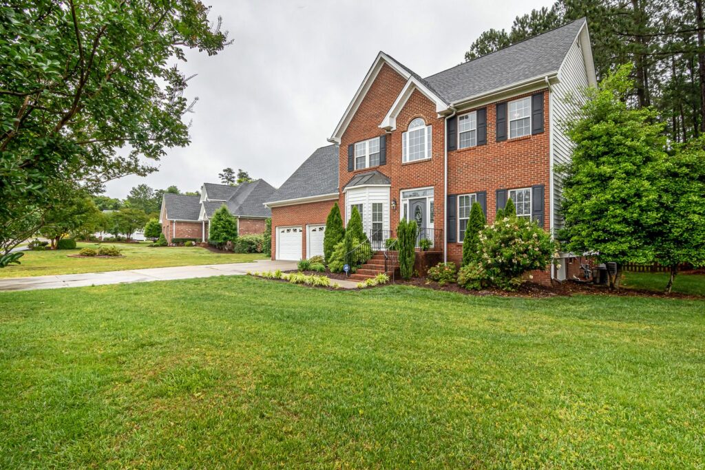 Two-story brick house with a mowed front lawn, foundation shrubs, and mature trees