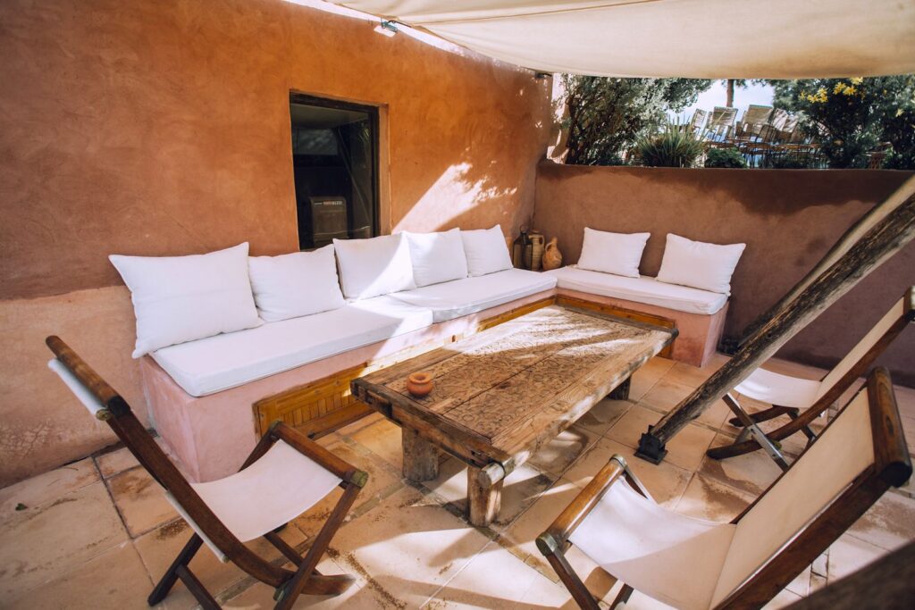 Built-in patio bench with white cushions under a shade sail, with a rustic table and sling chairs