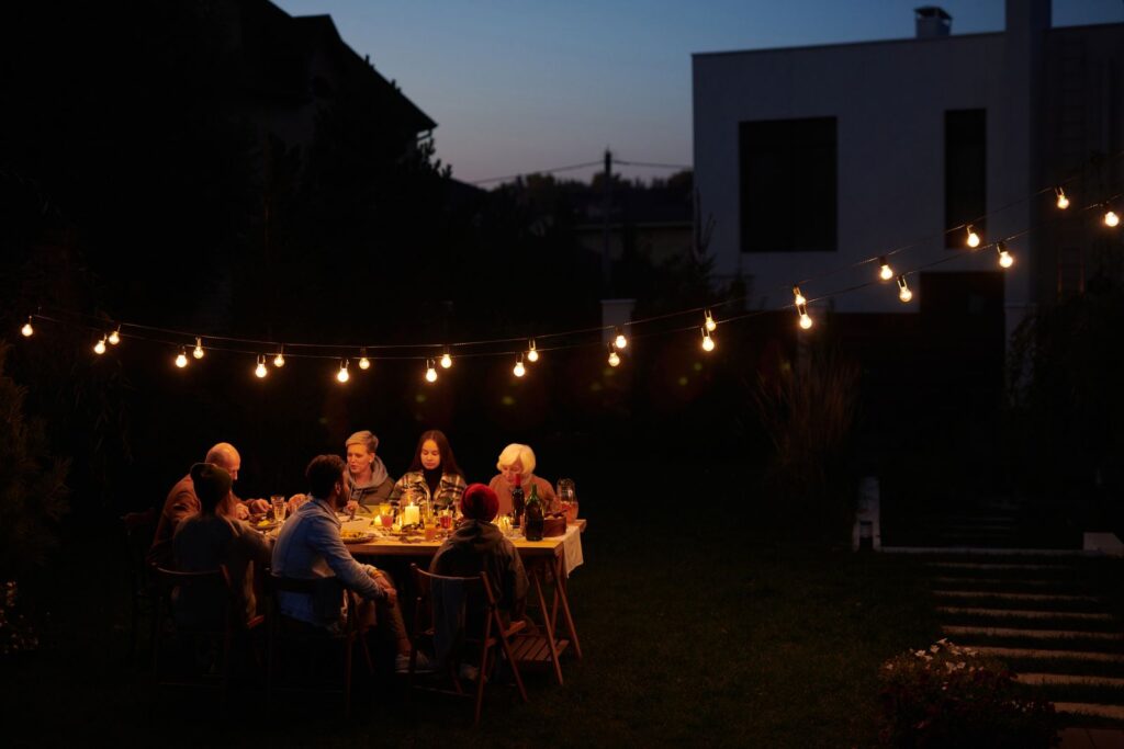 A family eating dinner outdoors at dusk under a canopy of string lights