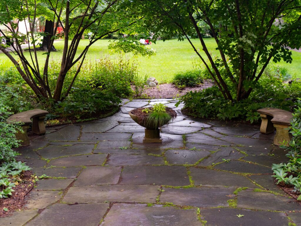 A flagstone patio with stone benches and a center planter, surrounded by shrubs and trees