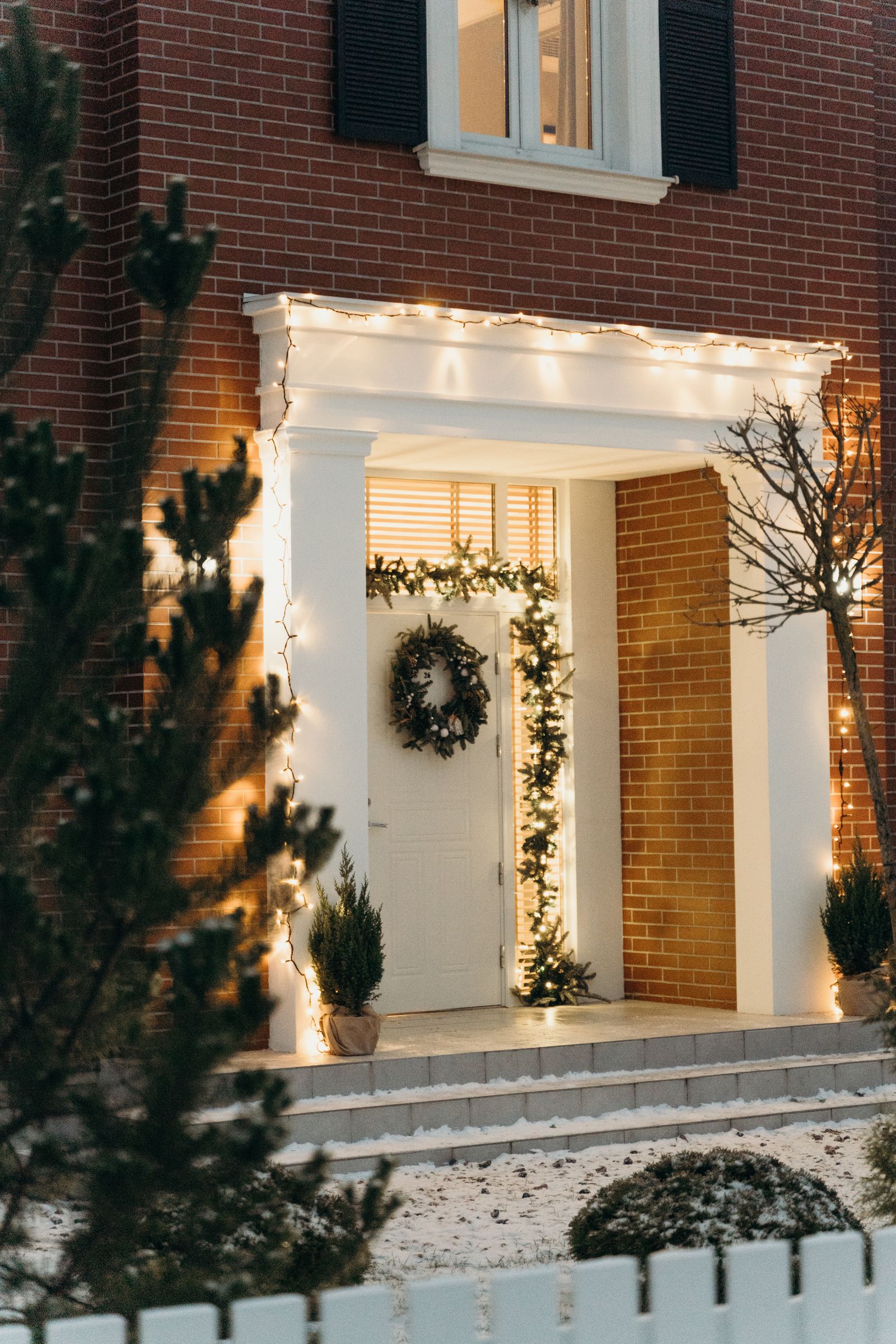 A brick front entrance decorated with a wreath, garland, string lights, and potted evergreens