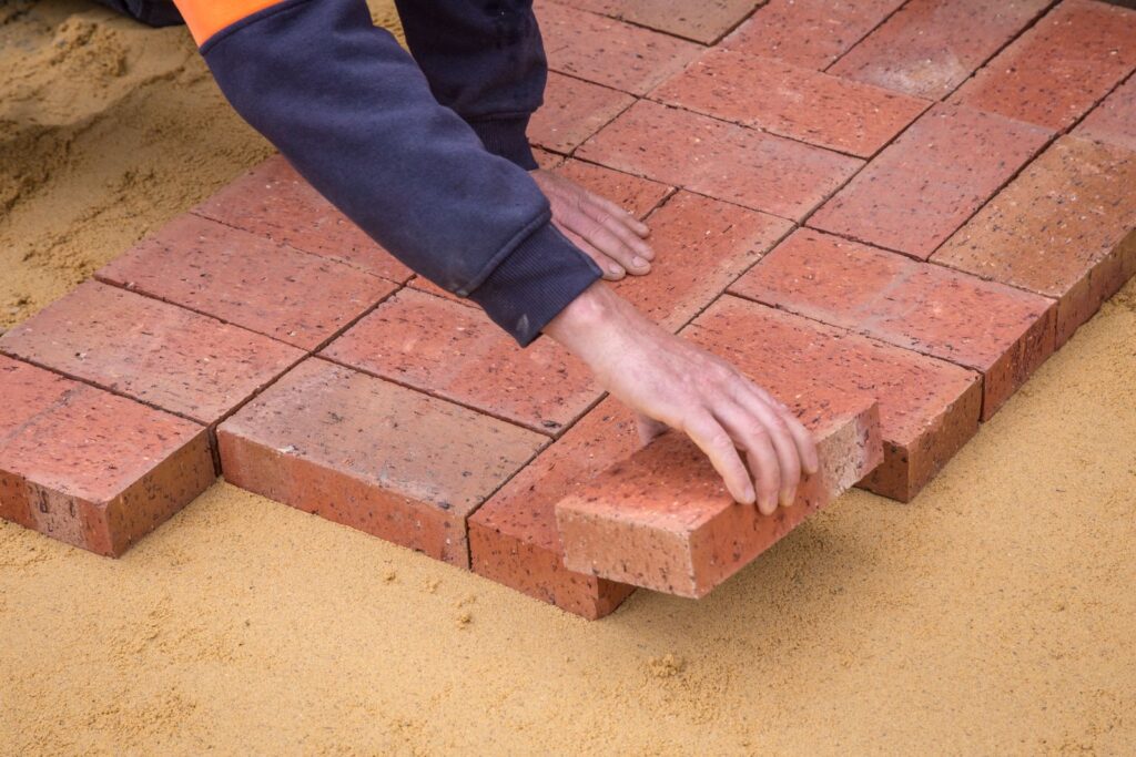 Hands laying red brick pavers on a bed of sand for a DIY path