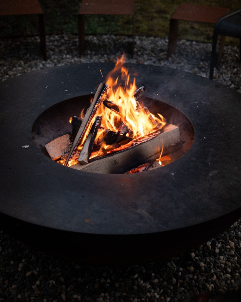 A wood fire burning in a wide steel fire bowl set on gravel, with chairs behind