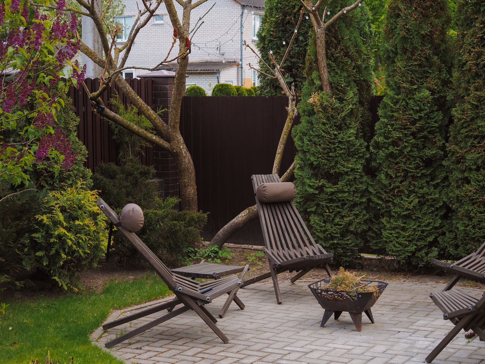 Wooden lounge chairs on a small paver patio beside a lawn and evergreen shrubs