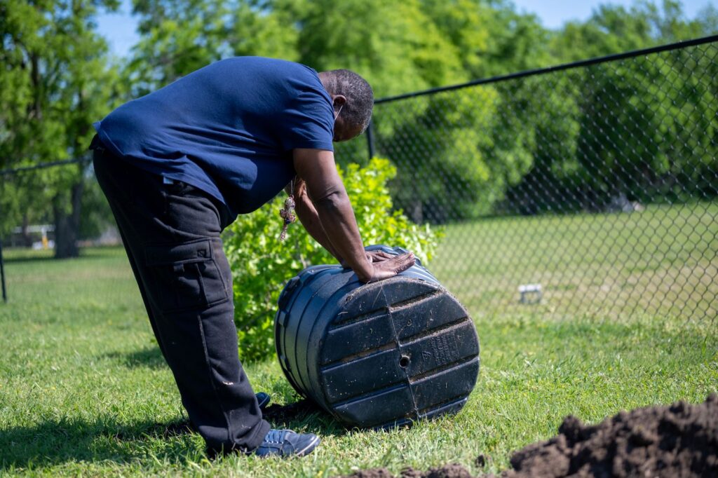 A man rolling a black plastic compost bin across a lawn to mix the contents