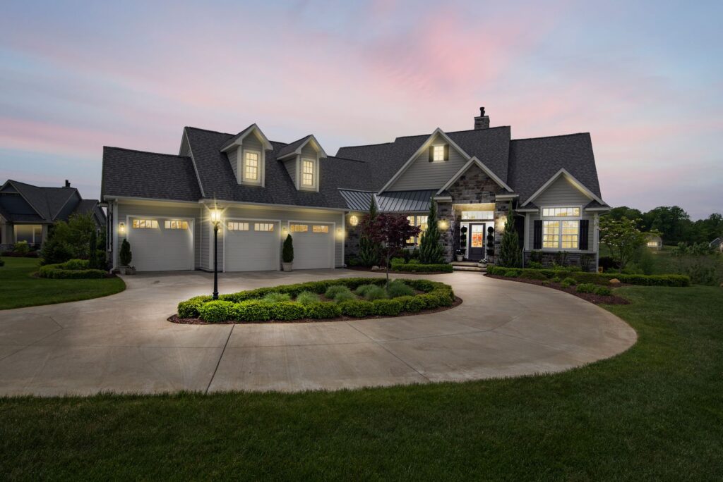 A suburban house at dusk with a lit driveway lamp, entry lights, and planted island bed