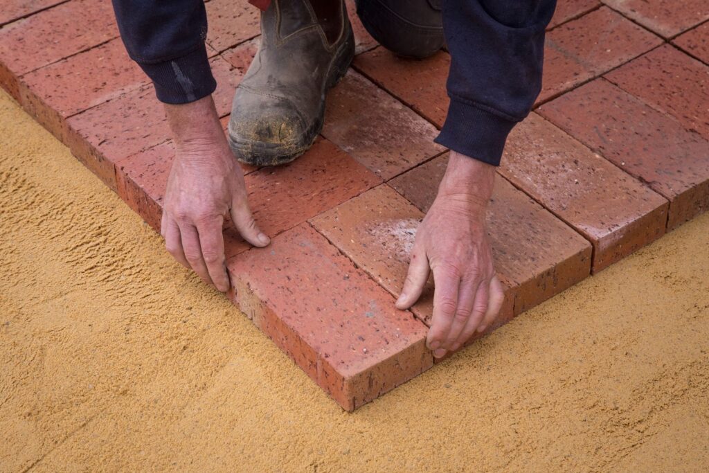 Hands setting brick pavers onto a screeded sand bed